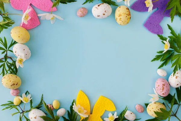ハッピーイースター カラフルなイースターバニー チョコレートの卵と青の背景に春の花のフレームは テキストのためのスペースとフラットレイアウト ピンクと黄色の人工的な装飾と新鮮な花 — ストック写真