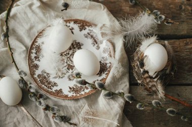 Rustic Easter still life. Natural egg in nest with feathers, vintage plate,  pussy willow branches and napkin on aged wood top view. Easter table decoration. Moody image