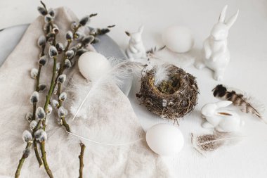 Stylish Easter still life. Natural easter eggs, nest, bunny, pussy willow branches, feathers, modern plate with napkin on white wooden table. Modern Easter table decoration