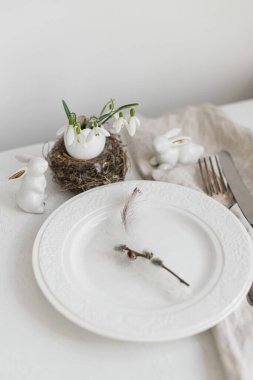 Pussy willow branch, feather on modern plate, napkin, cutlery, bunny and egg with snowdrops on white wooden table. Stylish Easter table setting. Modern Easter table decoration