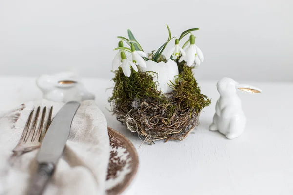 スタイリッシュなイースターテーブルの設定 巣の中の雪滴 現代のプレート上のナプキンの羽 カトラリー ウサギと白い木製のテーブルの上に天然の卵 現代イースターテーブル素朴な装飾 — ストック写真