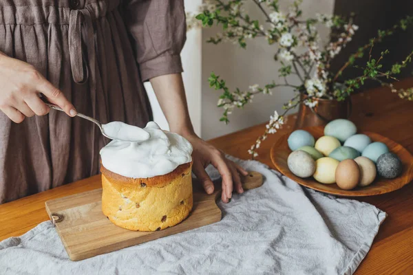 Woman decorating homemade easter bread with sugar glaze on rustic table with natural dyed eggs and spring blossom in room. Happy Easter! Woman baking stylish easter cake