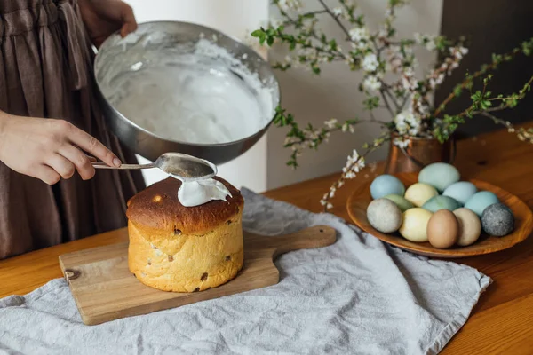 Woman decorating homemade easter bread with sugar glaze on rustic table with natural dyed eggs and spring blossom in room. Happy Easter! Woman baking stylish easter cake