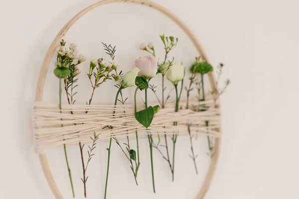 Stylish spring wreath with beautiful fresh flowers. Wooden hoop with flowers and thread hanging on white wall. Modern and creative floral handmade decor