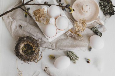 Paskalya kırsalı düzlüğü. Doğal yumurtalar, tüyler, söğüt dalları, yaşlı beyaz masada yuva ve mum. Pastel beyaz ve bej renkli kırsal Paskalya 'da hayat devam ediyor. Modern basit estetik.