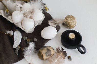 Tepside doğal paskalya yumurtaları, tüyler, söğüt dalları, beyaz ahşap masadaki keten peçeteye yuva. Paskalya kırsalında hayat. Pastel kahverengi renklerde basit Paskalya estetiği. Mutlu Paskalyalar