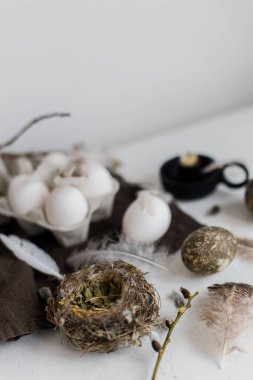 Paskalya kırsalında hayat. Tepside doğal paskalya yumurtaları, tüyler, söğüt dalları, beyaz ahşap masadaki keten peçeteye yuva. Pastel kahverengi renklerde basit Paskalya estetiği. Mutlu Paskalyalar