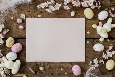 Paskalya tebrik kartı modeli, kırsal düzlük. Boş kart, renkli paskalya şekeri yumurtaları, tüyler, yıllanmış odunların üzerinde kiraz dalları. Mutlu Paskalyalar! Metin için boşluk içeren kağıt kart şablonu