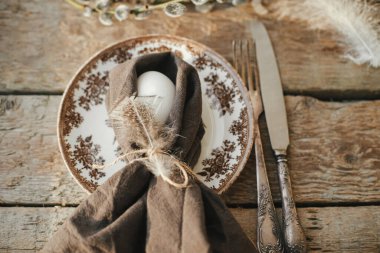 Rustic Easter table setting top view. Natural egg in napkin with feather, setting  with vintage plate, cutlery, pussy willow branches on aged wooden table. Easter table decoration