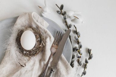 Stylish Easter table setting flat lay. Natural easter egg in nest, pussy willow branches, feathers on modern plate with napkin and cutlery on white wooden table. Modern Easter table decoration