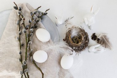 Stylish Easter flat lay. Natural easter eggs, nest, bunny, pussy willow branches, feathers, modern plate with napkin on white wooden table. Modern Easter table decoration, still life