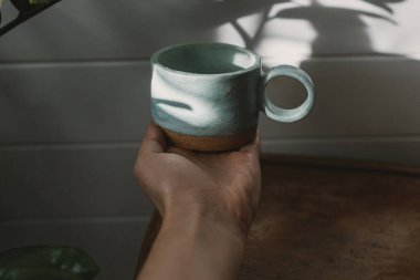 Modern kupa kadın elinde, güneş ışığında. Güneşli boho odasında çiçekli seramik minimalist bardakta el ele tutuşmak. Sabah kahvesi estetiği.