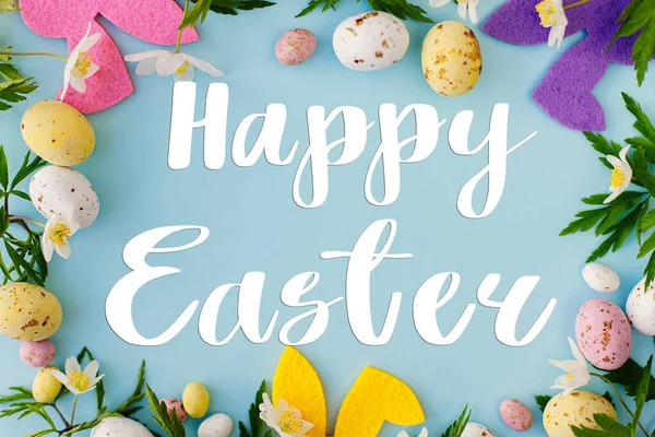Mutlu Paskalya tebrik kartları. Mutlu Paskalya metinleri, renkli tavşanlar, çikolatalı yumurtalar ve mavi arka planda bahar çiçekleri çerçevesi, düz yatak. Mevsimlik tebrik kartı, el yazması mektuplar.