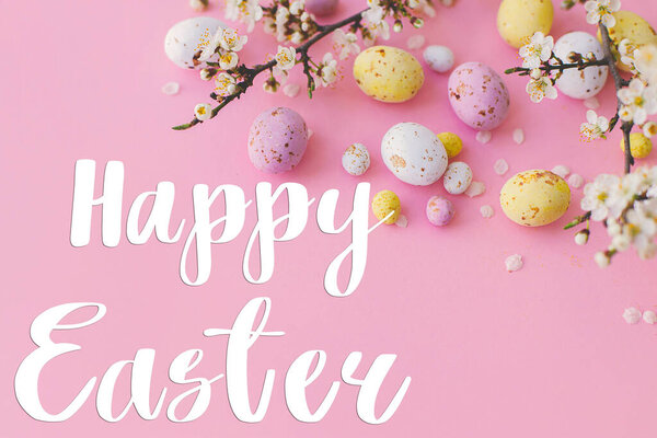 Happy Easter text and colorful Easter chocolate eggs and cherry blossoms border on pink background. Happy Easter greeting card. Seasons greeting card, handwritten lettering