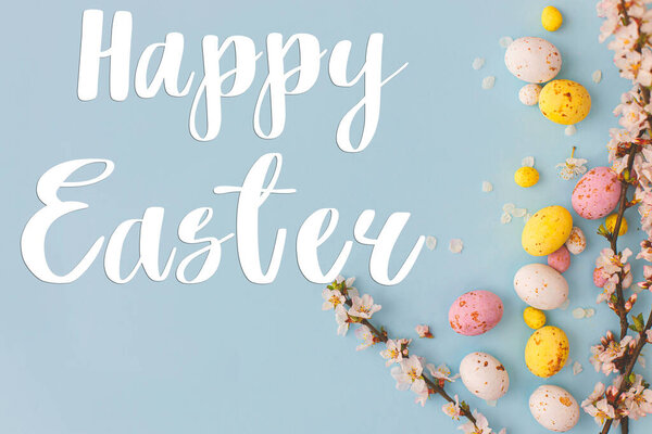 Happy Easter greeting card. Happy Easter text and colorful Easter chocolate eggs and cherry blossoms border on blue background flat lay. Seasons greeting card, handwritten lettering