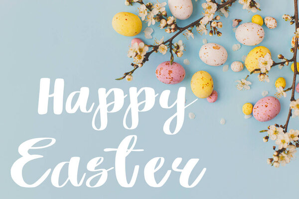 Happy Easter text and colorful Easter chocolate eggs and cherry blossoms border on blue background flat lay. Happy Easter greeting card. Seasons greeting card, handwritten lettering