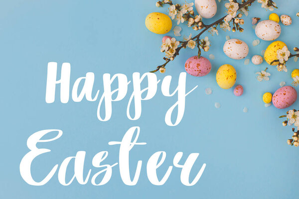 Happy Easter greeting card. Happy Easter text and colorful Easter chocolate eggs and cherry blossoms border on blue background flat lay. Seasons greeting card, handwritten lettering