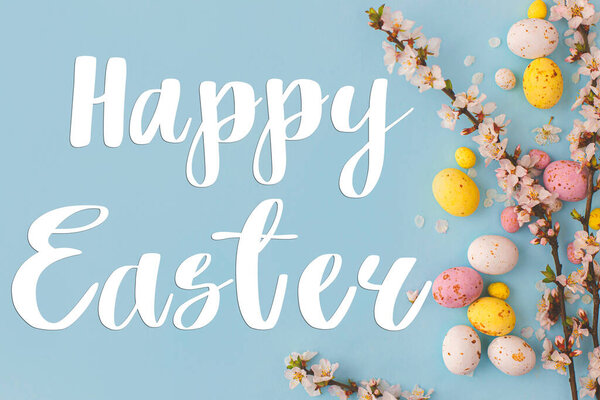 Happy Easter text and colorful Easter chocolate eggs and cherry blossoms border on blue background flat lay. Happy Easter greeting card. Seasons greeting card, handwritten lettering