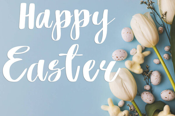 Happy Easter greeting card. Happy Easter text and modern stylish easter eggs, tulips and bunnies flat lay on blue background. Seasons greeting card, handwritten lettering