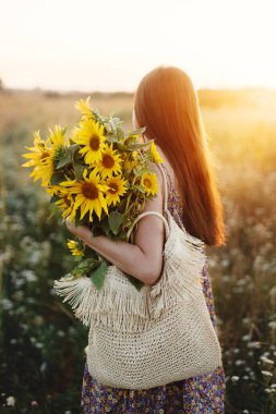 Çiçekli elbiseli şık genç bir kadın yaz tarlasında gün batımında ayçiçekleriyle yürüyor. Kırsal kesimde sakin bir atmosfer anı. Akşam çayırında elinde ayçiçekleri tutan güzel bir kadın
