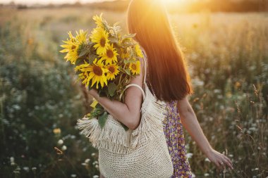 Çayırda gün batımının sıcacık ışığında ayçiçeği tutan güzel bir kadın. Kırsal kesimde sakin bir atmosfer anı. Çiçekli elbiseli şık bir bayan yaz akşamı tarlasında ayçiçekleriyle yürüyor.