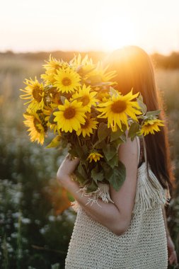 Çiçekli elbiseli şık genç bir kadın yaz tarlasında gün batımında ayçiçekleriyle yürüyor. Kırsal kesimde sakin bir atmosfer anı. Akşam çayırında elinde ayçiçekleri tutan güzel bir kadın