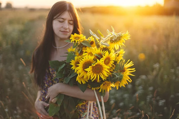 Çiçekli elbiseli şık genç bir kadın yaz tarlasında gün batımında ayçiçekleriyle yürüyor. Kırsal kesimde sakin bir atmosfer anı. Akşam çayırında elinde ayçiçekleri tutan güzel bir kadın