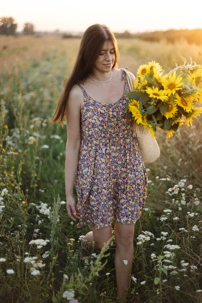 美しい女性は夏の牧草地で暖かい日没の光の中でひまわりを集める 田舎の静かな雰囲気の瞬間 夜のフィールドでひまわりを選ぶ花のドレスでスタイリッシュな若い女性 — ストック写真