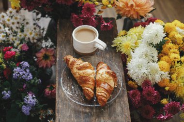 Sıcak bir fincan kahve ve yeni pişmiş kruvasan. Kırsal ahşap arka planda ve güzel sonbahar çiçekleri. İyi sabahlar. Kapuçino ve kruvasan tabaklı şık bir kupa.