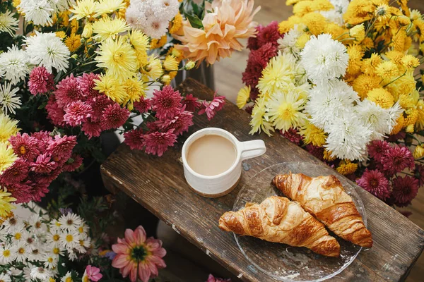 Sıcak bir fincan kahve ve yeni pişmiş kruvasan. Kırsal ahşap arka planda ve güzel sonbahar çiçekleri. İyi sabahlar. Kapuçino ve kruvasan tabaklı şık bir kupa.