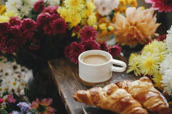 Sıcak bir fincan kahve ve yeni pişmiş kruvasan. Kırsal ahşap arka planda ve güzel sonbahar çiçekleri. İyi sabahlar. Kapuçino ve kruvasan kaplı şık bir kupa, kasvetli bir görüntü.