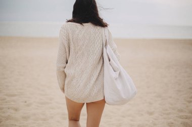 Rüzgarlı saçları ve çantasıyla kumlu sahilden denize yürüyen zarif bir kadın. Kaygısız bir an. Örgü kazaklı şık genç bir bayan tatilin tadını çıkarıyor ve sahilde dinleniyor.