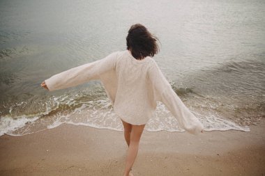 Kumsal ve deniz dalgaları, arka planda bulanık, kaygısız, örgü süveter giymiş kumsalda yürüyen güzel bir kadın görüntüsü. Yaz tatili kavramı. Mutlu kadın sahilde tatilin tadını çıkarıyor.