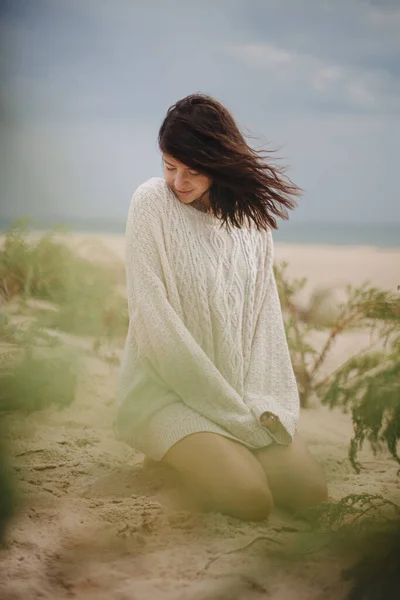 Beautiful woman with windy hair sitting on sandy beach on background of green grass and sea, calm tranquil moment. Stylish young female in knitted sweater enjoying  and relaxing on coast