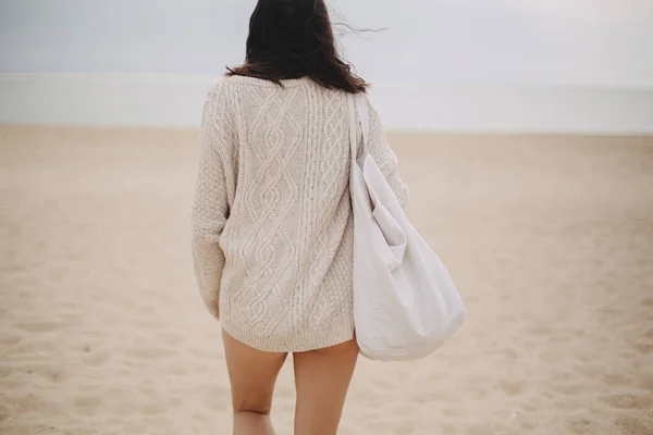 Beautiful stylish woman with windy hair and tote bag walking on sandy beach to sea, carefree moment. Stylish young female in knitted sweater enjoying vacation and relaxing on coast