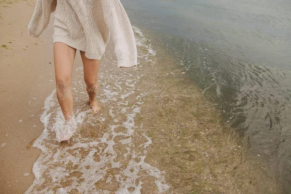 Kaygısız güzel kadın kumlu sahilde çıplak ayakla koşuyor soğuk deniz dalgalarıyla, eğleniyor. Kesik manzara. Şık, mutlu bir bayan, dinleniyor ve sahilde tatilin tadını çıkarıyor. Su sıçratan dalgalarda ayaklar