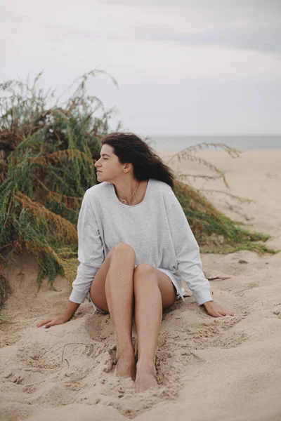 Rüzgarlı saçlı, kumlu sahilde yeşil çimlerin ve denizin arka planında oturan güzel bir kadın, sakin bir an. Kıyıda dinlenen zarif genç bir bayan. Tatil havası.