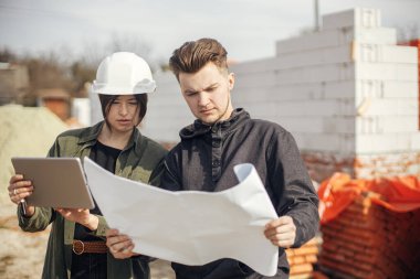 Stylish woman architect with tablet  and foreman checking blueprints at construction site. Young engineer and construction workers in hardhat looking at plans of new modern house. Teamwork