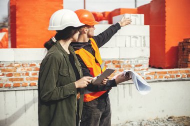 Tabletli şık bir kadın mimar ve ustabaşı inşaat alanının planlarını kontrol ediyor. Genç mühendis ve inşaat işçileri yeni modern evin planlarına bakıyorlar. Takım çalışması.