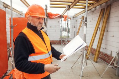 Senior male foreman in hardhat checking blueprints of building new modern house. Man engineer or construction worker looking at plans at construction site. Copy space