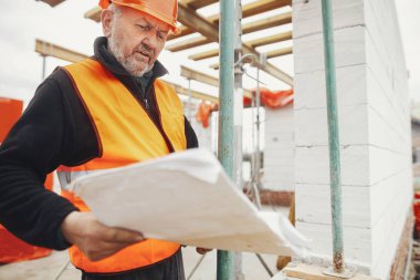 Senior man engineer or construction worker in hardhat looking at blueprints at building new modern house. Male architect or contractor checking plans at construction site. Copy space