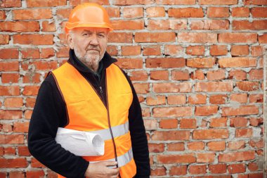 Senior man engineer or construction worker in hardhat standing with blueprints at masonry brick wall at building new house. Portrait of male foreman holding plans at construction site
