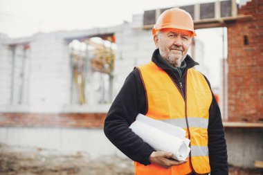 Senior man engineer or construction worker in hardhat standing with  blueprints at building new modern house. Male architect or contractor checking plans at construction site. Copy space
