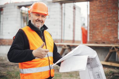 Senior male foreman in hardhat approving building new modern house according to blueprints. Portrait of happy man engineer or construction worker with plans at construction site