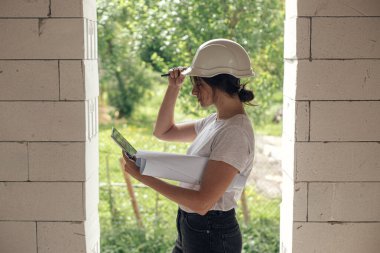 İnşaat alanındaki dijital planlara bakan sert şapkalı şık kadın mühendis. Tabletli genç bayan mimar yeni modern evdeki pencerelerle planları karşılaştırıyor.