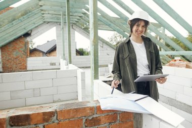 Tabletle şık bir kadın mimar ya da mühendis modern çiftlik evinin ahşap çatı çerçevesiyle ilgili planları kontrol ediyor. İnşaat alanındaki dijital planlara bakan kasklı mutlu genç bir kadın.