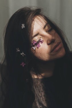 Yüzünde leylak yaprakları ve gün ışığında saçları olan güzel, şehvetli bir kadın. Genç esmerin karamsar portresi. Yüzünde çiçekler ve gölgeler var. Deri bakımı ve doğal kozmetik konsepti