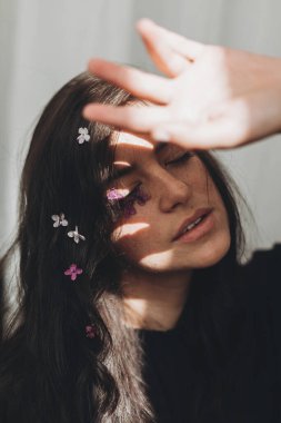 Yüzünde leylak yaprakları ve güneş ışığında poz veren güzel, şehvetli bir kadın. Kadınlık. Genç esmerin karamsar portresi. Yüzünde çiçekler ve gölgeler var. Cilt bakımı ve kişisel bakım