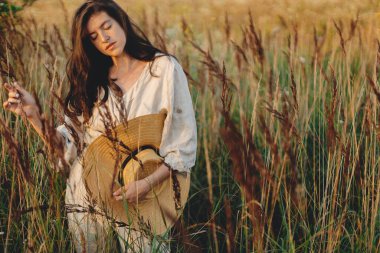 Akşamları yabani otların arasında poz veren hasır şapkalı şık boho kadın. Yaz tatili ve seyahat. Kırsal keten kumaş içinde kaygısız genç bir kadın yaz çayırlarında dinleniyor. Atmosferik an