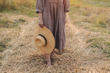 Gün batımında saman tarlasında çıplak ayakla yürüyen şapkalı kadın, yaklaş. Kırsal yaşam. Atmosferik sükunet anı. Kırsal kesimde, keten elbiseli genç bir kadın dinleniyor.